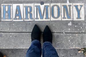 Looking down at a person's lower legs and boots standing at tiles for a street marker that says Harmony 