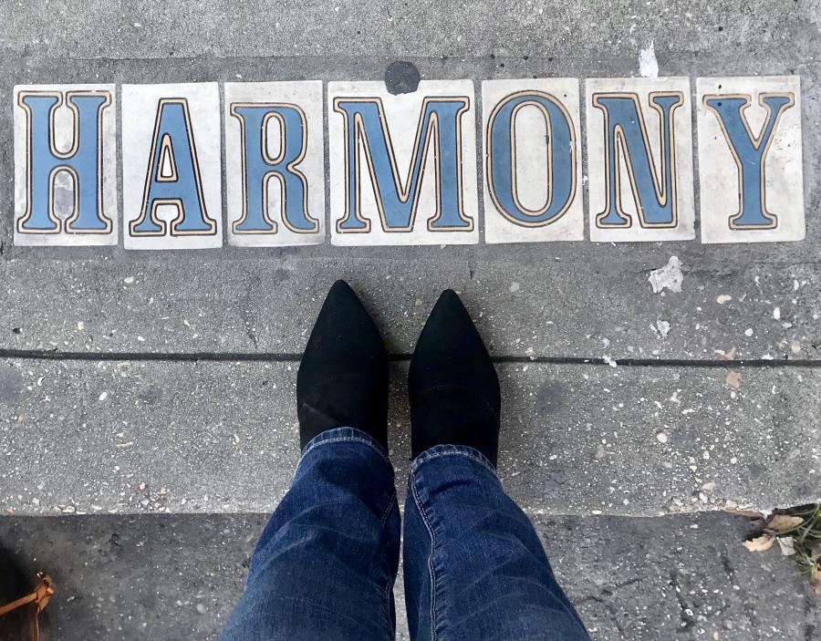 Looking down at a person's lower legs and boots standing at tiles for a street marker that says Harmony 